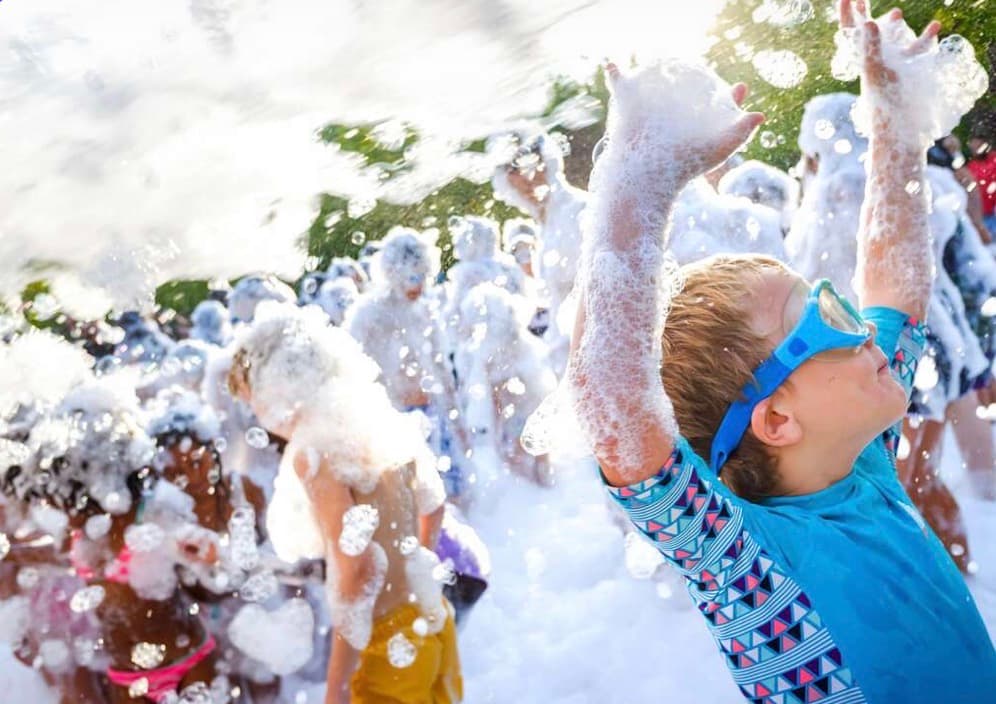 Boy in blue goggles raises foam-covered arms at a sunny outdoor foam party.