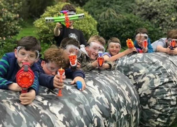 Boys wearing safety goggles aim toy blasters from behind large camouflage inflatable bunkers outdoors.