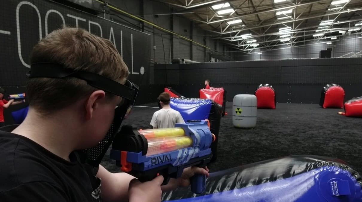 Boy wearing a mask aims a blue toy blaster in an indoor obstacle arena.