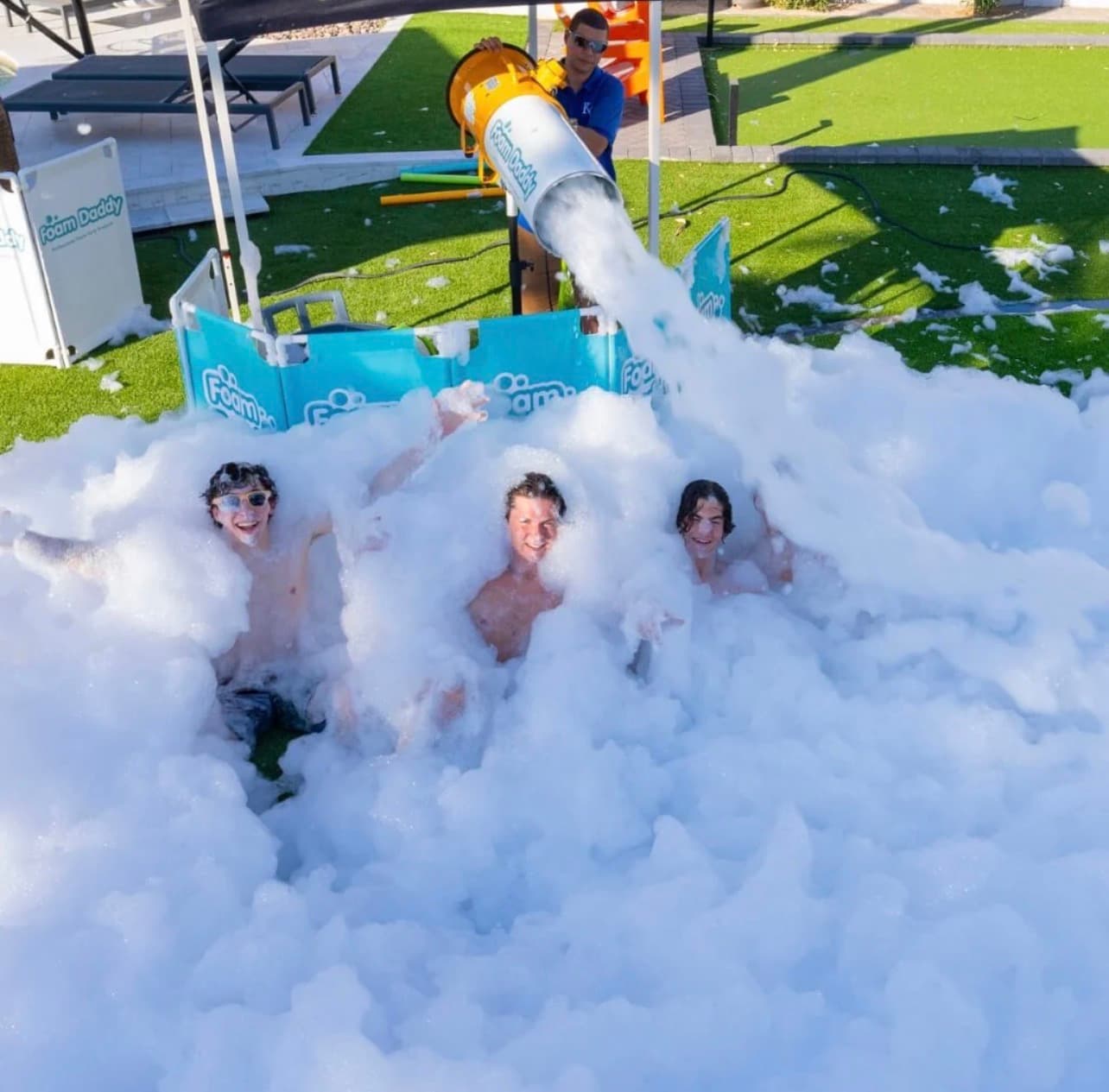 Three men laugh while being sprayed with thick white foam from a large foam cannon.