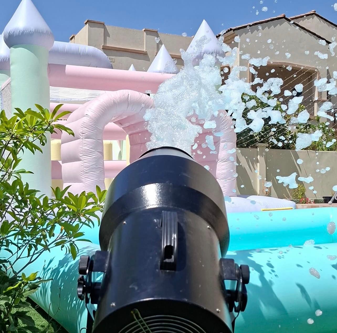 Black foam cannon spraying white foam toward a pastel-colored inflatable bounce house in a backyard.