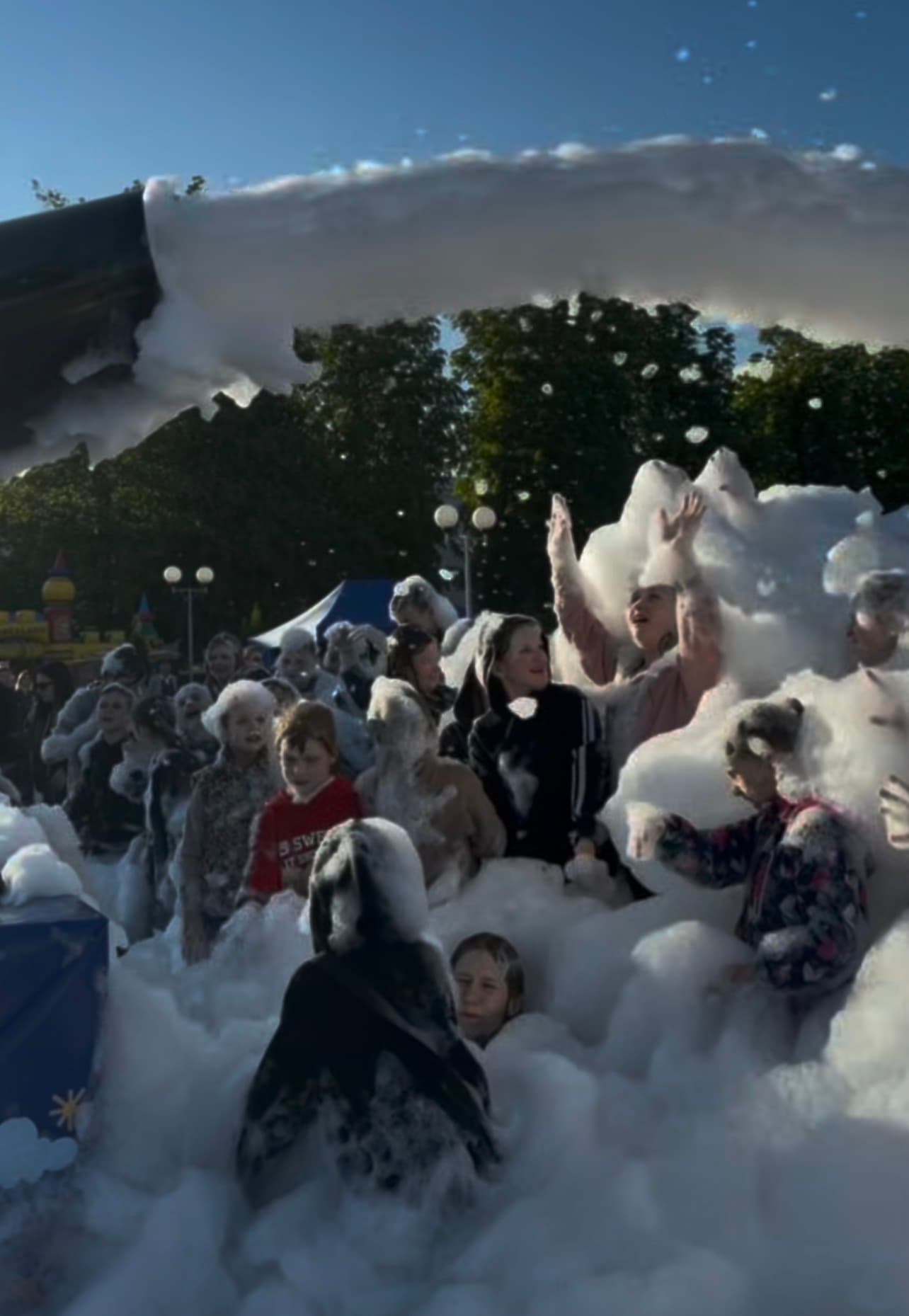 Excited children and adults play in thick white foam sprayed from a cannon outdoors.