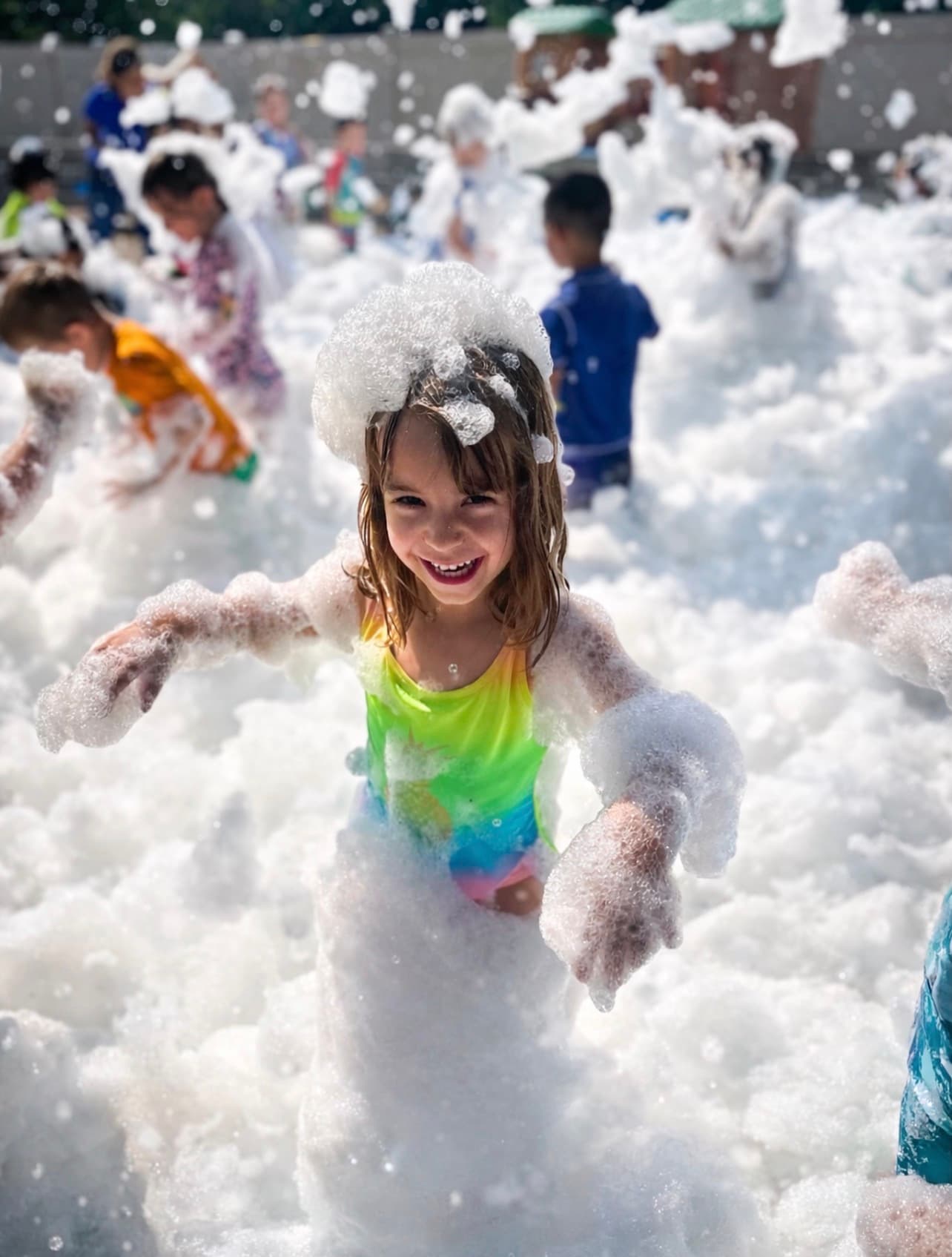 Smiling girl in a rainbow swimsuit covered in white foam at an outdoor foam party.